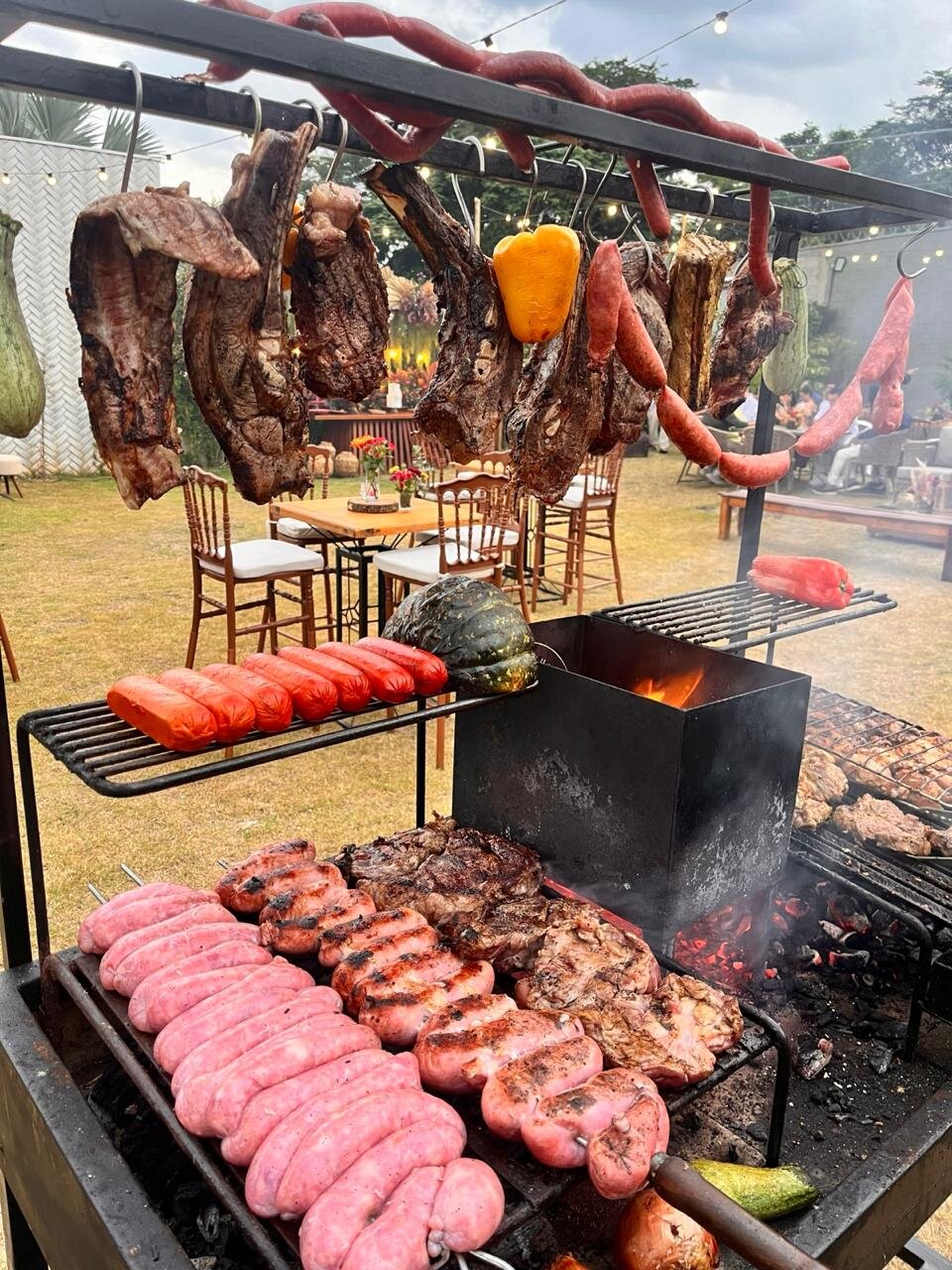 Parrilla e trempe com carnes e linguiças montadas em casamento, com as mesas dos convidados ao fundo